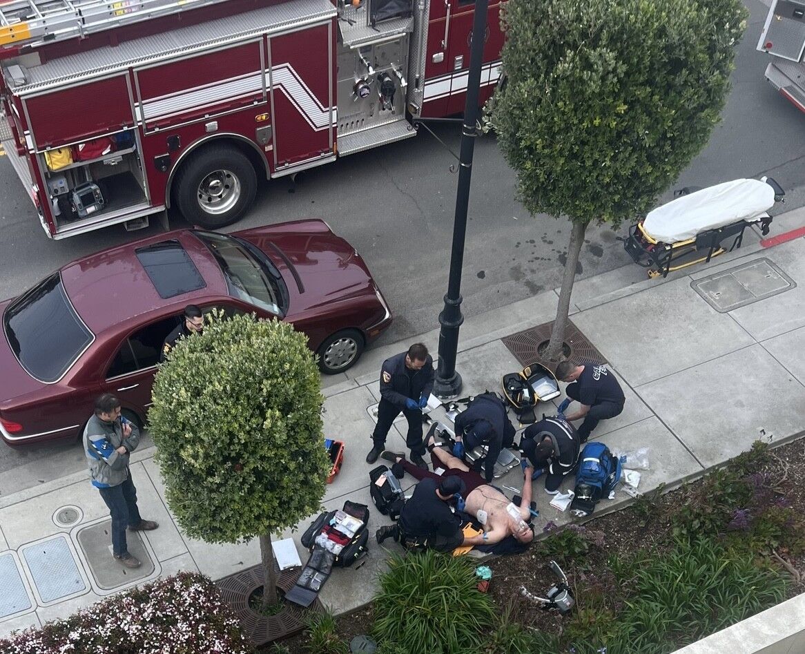 Matthew Burrows experienced cardiac arrest in Half Moon Bay in 2023. His initial rescuer, Peter Williams, stands at left while an emergency medical team uses a defibrillator on Burrows.